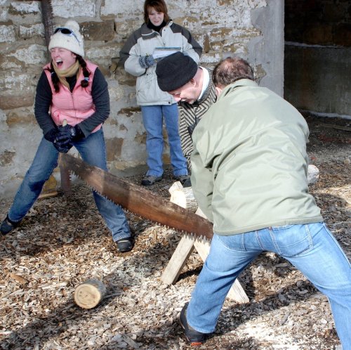 Two people saw a log together while a third person watches, smiling, in a rustic outdoor setting.