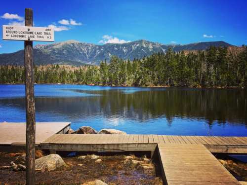 A wooden dock extends into a calm lake, surrounded by trees and mountains under a clear blue sky. Trail sign visible.