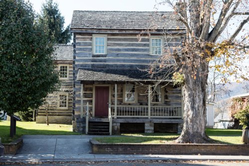 A rustic log cabin with a porch, surrounded by trees and grass, on a sunny day.