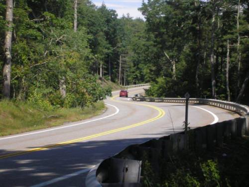 A winding road curves through a forested area, with a red car driving along the path.
