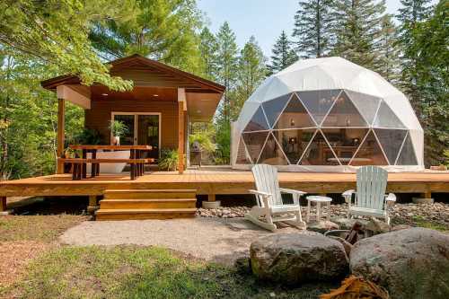 A cozy wooden cabin and a geodesic dome on a deck surrounded by trees, with two white chairs in the foreground.