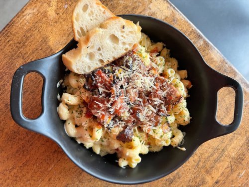 A black bowl filled with creamy macaroni and cheese topped with crispy bacon and grated cheese, served with a slice of bread.
