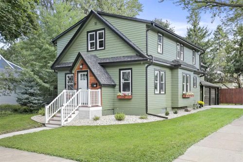 A green two-story house with white trim, a front porch, and flower boxes, surrounded by a well-maintained lawn.