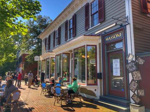A charming café with outdoor seating, surrounded by trees and a sunny blue sky, inviting patrons to enjoy their meals.