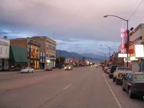 A quiet street scene at dusk, featuring shops, parked cars, and mountains in the background under a cloudy sky.