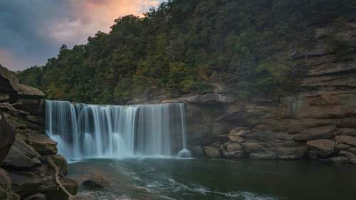 A serene waterfall cascades over rocks into a calm pool, surrounded by lush greenery and a cloudy sky.