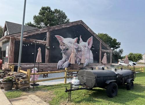 A mural of two pigs on a building, with a barbecue smoker in front and outdoor seating under umbrellas.