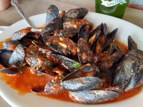A plate of mussels in a rich tomato sauce, garnished with herbs, served with a drink in the background.