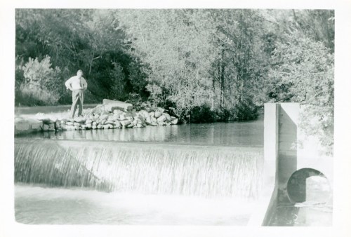A black and white photo of a man standing by a river with a small waterfall and trees in the background.