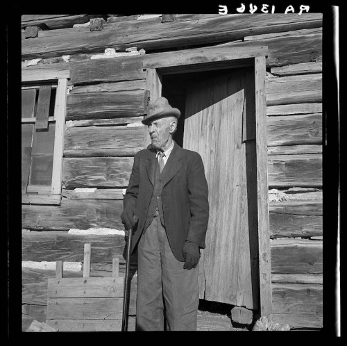 An elderly man in a suit and hat stands by a wooden cabin door, holding a cane.