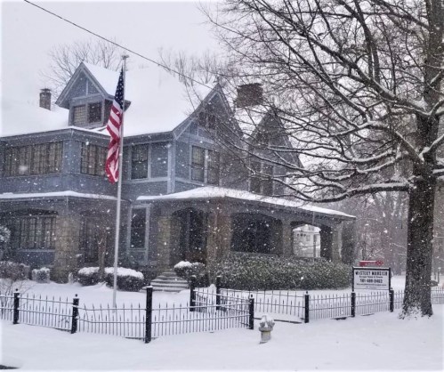 A large blue house covered in snow, with an American flag and a "For Sale" sign in front. Snowflakes are falling.