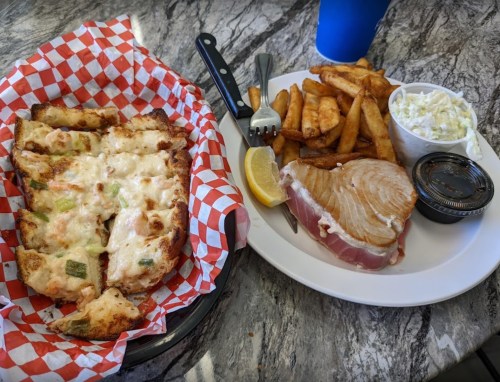 A plate with grilled fish, fries, coleslaw, and a lemon wedge, alongside a basket of cheesy bread.