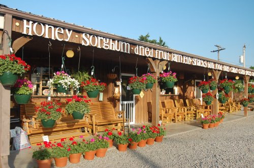 A rustic storefront with hanging flower baskets, wooden rocking chairs, and potted plants, showcasing local products.
