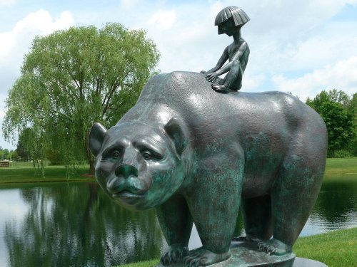 A bronze sculpture of a bear with a child sitting on its back, set by a serene pond and lush greenery.