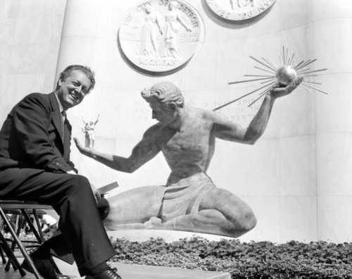 A man sits on a chair beside a large statue depicting a figure holding a globe and a sunburst, with a backdrop of medallions.