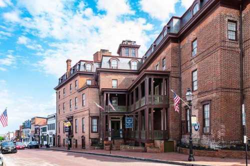 Historic brick building with a porch, flags, and a clear blue sky in the background, located on a street.