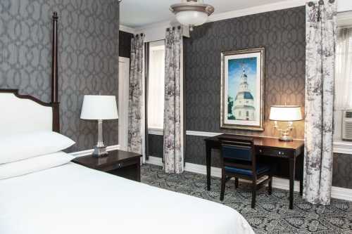 Cozy hotel room featuring a bed, desk, lamp, and patterned wallpaper with a framed painting on the wall.