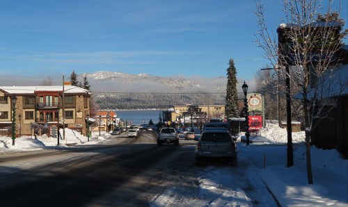Snow-covered street scene with buildings, parked cars, and a lake in the background under a clear blue sky.