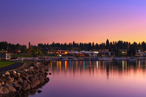 A serene lakeside view at dusk, with colorful lights reflecting on calm water and trees in the background.
