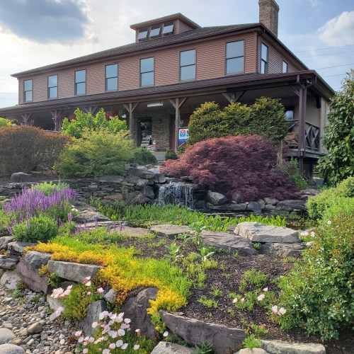 A charming two-story building surrounded by colorful gardens and a small waterfall, under a partly cloudy sky.