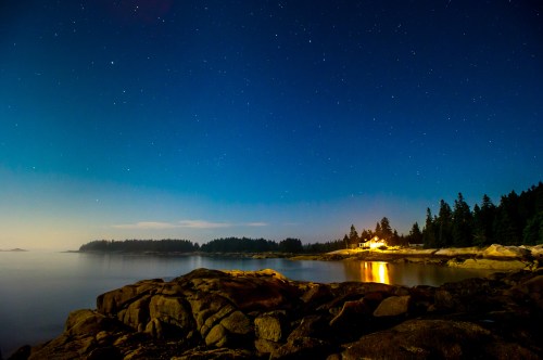A serene night scene of a rocky shoreline under a starry sky, with a distant house illuminated by warm light.