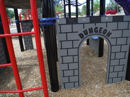 Playground structure featuring a "Dungeon" entrance with a brick design and colorful play equipment nearby.