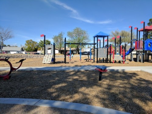 A playground with colorful equipment, including slides and climbing structures, surrounded by trees and a clear blue sky.