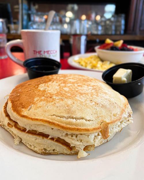 A stack of fluffy pancakes with butter and syrup, with a coffee cup and fruit bowl in the background.
