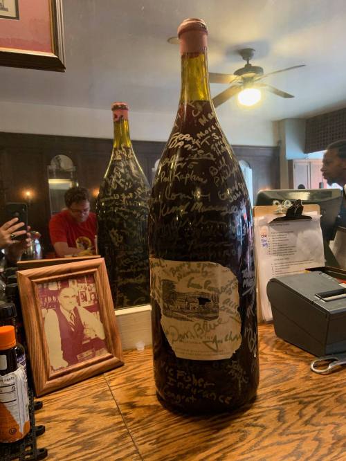 A large wine bottle with signatures on it, displayed on a wooden table in a bar setting, with a mirror reflecting the scene.