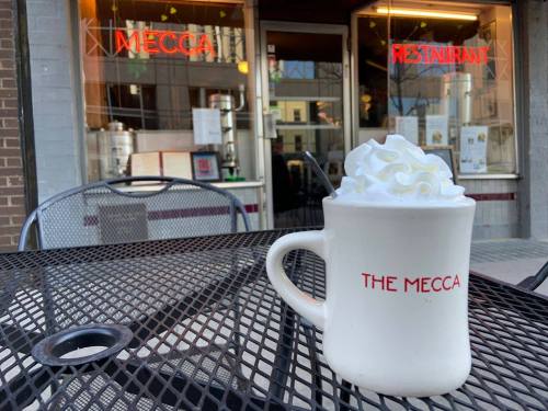 A white mug with "THE MECCA" printed on it, topped with whipped cream, sits on a black metal table outside a restaurant.