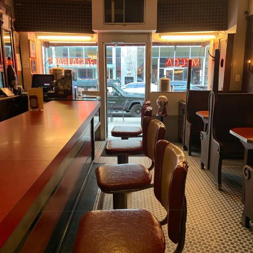 A diner interior with a long counter, empty chairs, and a view of the street through large windows.