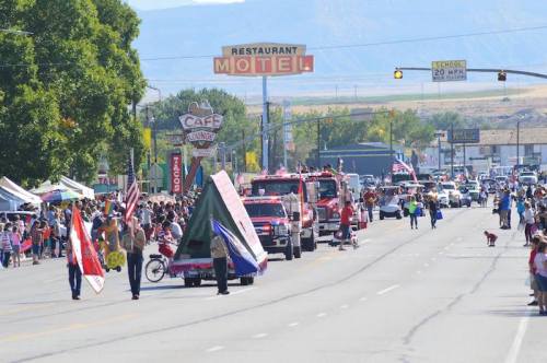 A lively parade with floats, flags, and fire trucks on a sunny street, surrounded by spectators and shops.