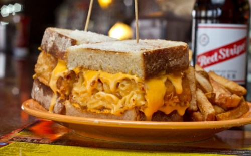 A hearty sandwich with macaroni and cheese, crispy chicken, and melted cheese, served with fries and a soda.