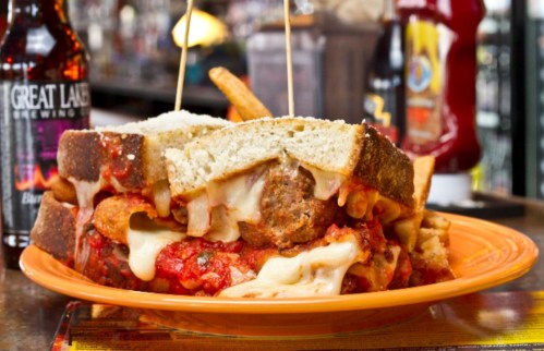 A hearty meatball sandwich with melted cheese, marinara sauce, and fries on an orange plate, set in a casual dining setting.