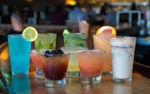 A colorful array of cocktails and drinks lined up on a bar, featuring various garnishes and glass styles.