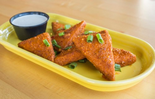 A yellow plate with triangular fried snacks topped with green onions, accompanied by a small cup of dipping sauce.