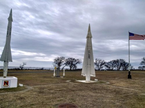 Three missile replicas stand in a grassy area, with an American flag waving in the background under a cloudy sky.