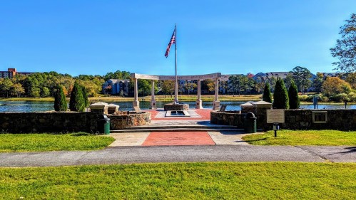 A serene park scene featuring a flagpole, stone walkway, and a lake surrounded by trees and buildings in the background.