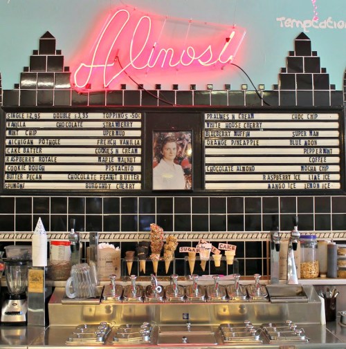 A retro ice cream shop counter with a neon sign, menu boards, and various toppings displayed.