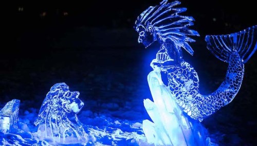 An illuminated ice sculpture of a mermaid and a lion, glowing blue against a dark background.