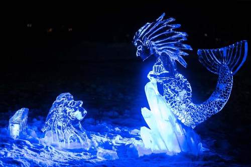 An illuminated ice sculpture of a mermaid facing a lion, surrounded by snow and blue light.