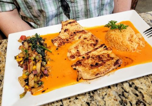 A plate of grilled fish with rice and a colorful vegetable medley, served on a white dish with a vibrant sauce.