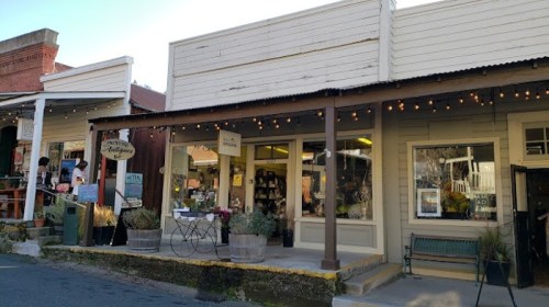A charming storefront with string lights, displaying various items, surrounded by rustic buildings in a quaint town.