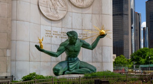 A green statue of a man holding a globe and a small figure, set against a stone wall with city buildings in the background.