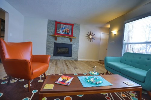 A cozy living room featuring an orange armchair, a teal sofa, a coffee table, and a stone fireplace with a colorful artwork above.