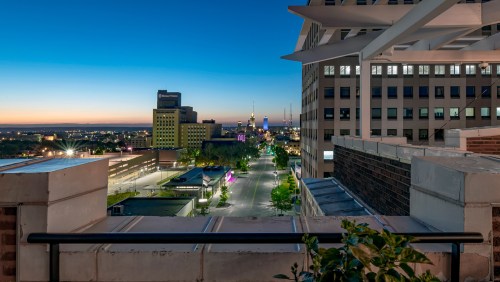 City skyline at dusk, featuring buildings and a clear sky transitioning from blue to orange. Street lights illuminate the road.