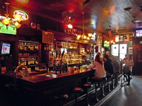 A cozy bar interior with a wooden counter, shelves of liquor, and patrons enjoying drinks in a warm, inviting atmosphere.