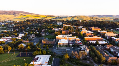 Aerial view of a university campus surrounded by green hills and fields, with buildings and trees visible.
