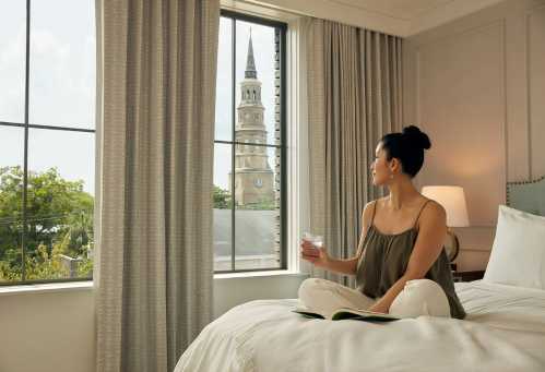 A woman sits on a bed, holding a drink and looking out a window at a church steeple in the distance.