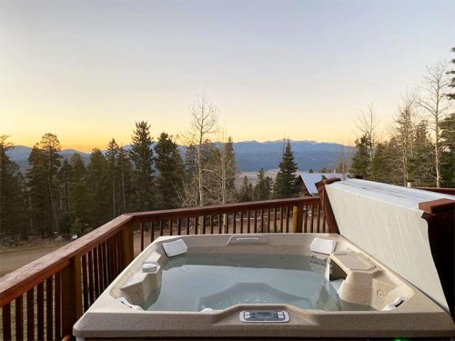 A hot tub on a wooden deck with a scenic view of mountains and trees at sunset.